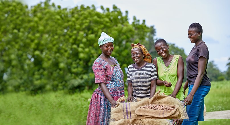 Four women standing in front of a sack of shea nuts, smiling and laughing